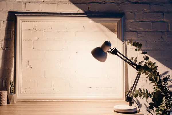 Lámpara de mesa iluminando pared de ladrillo blanco vacío con marco, concepto de maqueta - foto de stock