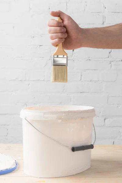 Cropped image of man holding brush in white paint above bucket — Stock Photo