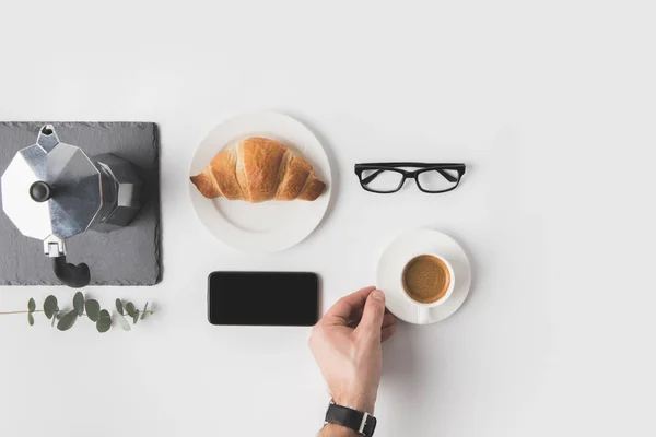 Schnappschuss eines Mannes, der eine Tasse Kaffee mit Smartphone, Brille und Croissant zum Frühstück in der Nähe auf weißer Fläche hält — Stockfoto