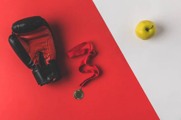 Вид сверху боксёрской перчатки с медалью и яблоком на красно-белой поверхности — стоковое фото