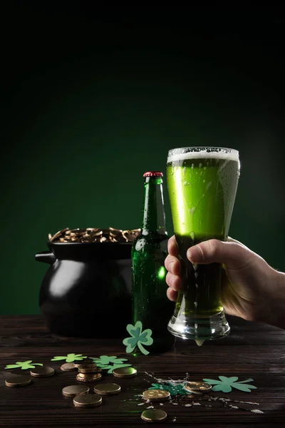 Imagen recortada del hombre sosteniendo un vaso de cerveza verde con una olla de monedas en la mesa, concepto de día de San Patricio - foto de stock