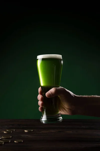 Обрезанный образ человека, держащего стакан зеленого пива над столом с монетами, День святого Патрика концепции — стоковое фото