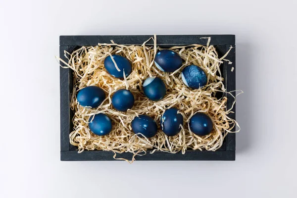 Top view of blue painted easter eggs in wooden box with decorative hay on white — Stock Photo