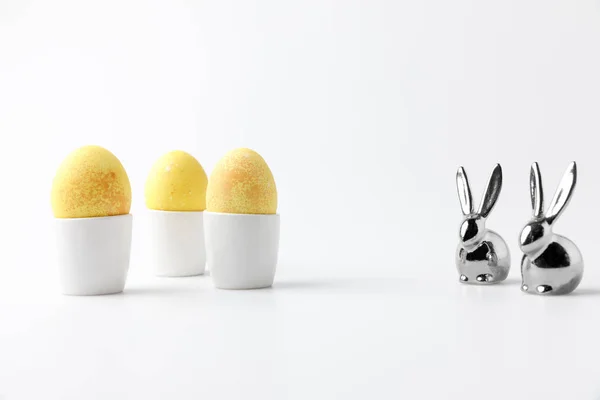 Желтые окрашенные пасхальные яйца в яичные стойки и статуэтки кроликов на белом — стоковое фото
