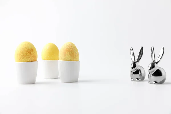 Huevos de Pascua pintados de amarillo en puestos de huevo y estatuillas de conejos sobre blanco - foto de stock