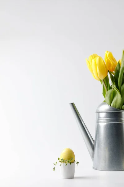 Huevo de Pascua pintado amarillo en soporte de huevo y tulipanes en regadera en blanco - foto de stock