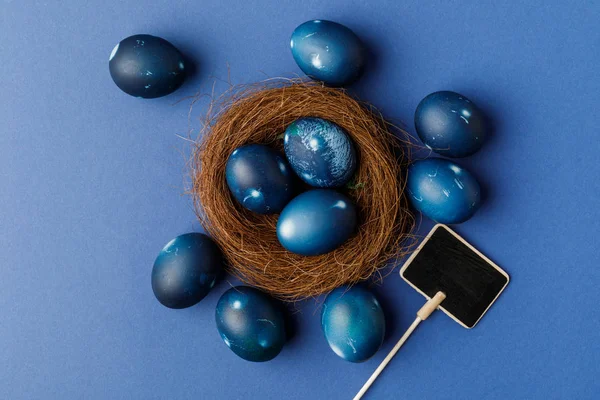 Vista superior de huevos de Pascua pintados de azul en nido decorativo sobre superficie azul - foto de stock