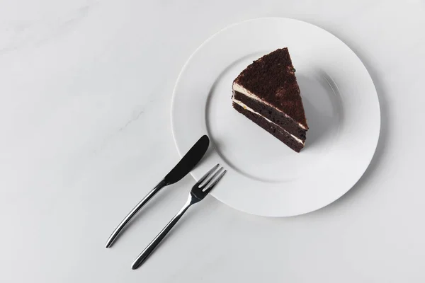 Pastel de chocolate en plato con tenedor y cuchillo en superficie blanca - foto de stock