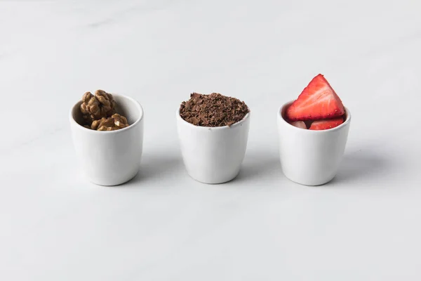 Крупный план трех чаш с грецкими орехами, тертым шоколадом и клубникой на сером — стоковое фото