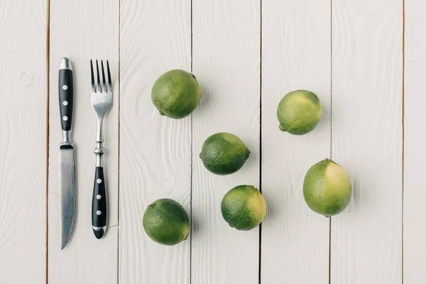 Top view of cutlery and green limes on white wooden background — Stock Photo
