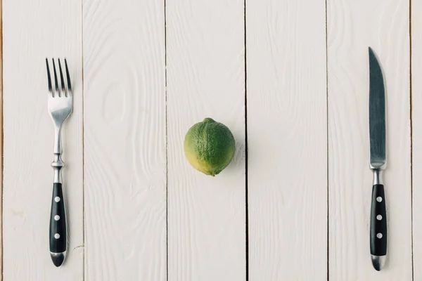 Top view of cutlery and green lime on white wooden background — Stock Photo