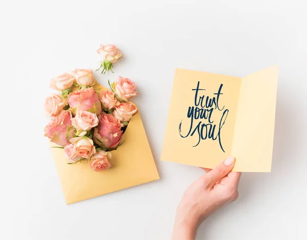Рука тримає папір зі знаком TRUST Your SOUL поруч з рожевими квітами в конверті ізольовані на білому — стокове фото