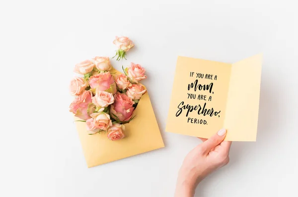 Hand holding paper with mothers day phrase beside pink flowers in envelope isolated on white — Stock Photo