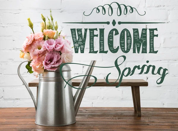 Hermosas flores florecientes en una regadera y un pequeño banco en una superficie de madera con letras WELCOME SPRING - foto de stock