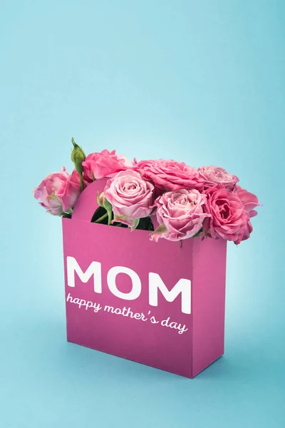 Nahaufnahme von schönen blühenden rosa Rosen in dekorativer Papiertüte mit glücklicher Muttertagsbeschriftung isoliert auf blauem Grund — Stockfoto
