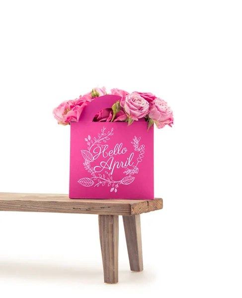 Vue rapprochée de fleurs roses à fleurs tendres en sac en papier rose avec lettrage HELLO APRIL sur banc en bois isolé sur blanc — Photo de stock