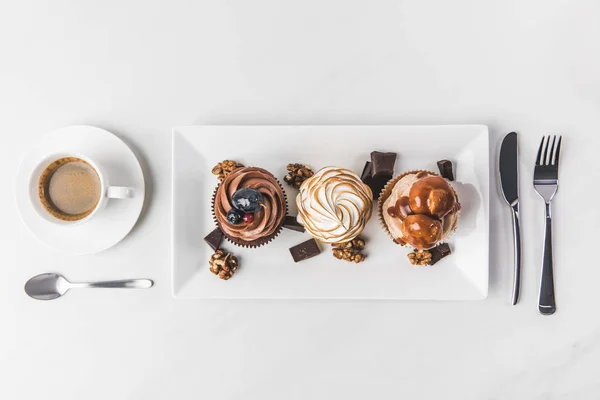 Vue de dessus de divers cupcakes sur assiette, tasse de café et couverts isolés sur blanc — Photo de stock