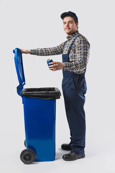 Side view of young cleaner in uniform with can standing at trash bin isolated on grey — Stock Photo