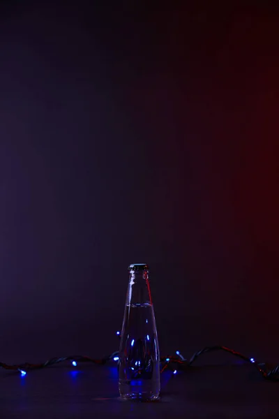 Bouteille d'eau fermée et guirlande bleue sur une surface sombre — Photo de stock