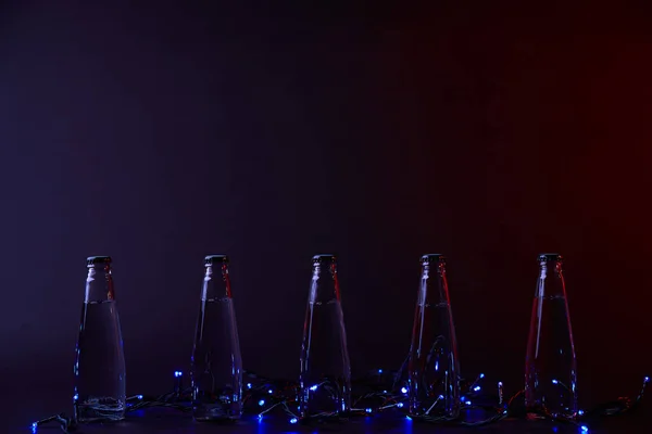 Закрытые бутылки воды с голубой гирляндой на темной поверхности — стоковое фото