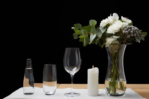Buquê em vaso com garrafa de água e copos vazios na mesa ao lado da vela no fundo preto — Fotografia de Stock