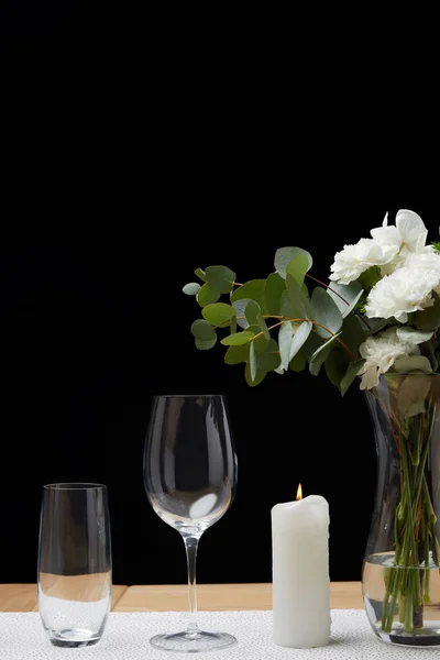 Flores en jarrón con vasos vacíos en la mesa junto a la vela sobre fondo negro - foto de stock