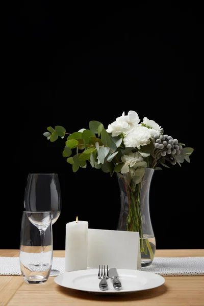 Dinnerware with glasses on table next to blank card and bouquet in vase on black background — Stock Photo