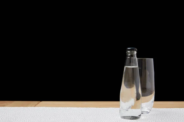 Vidrio y botella vacíos con agua sobre mantel sobre fondo negro - foto de stock