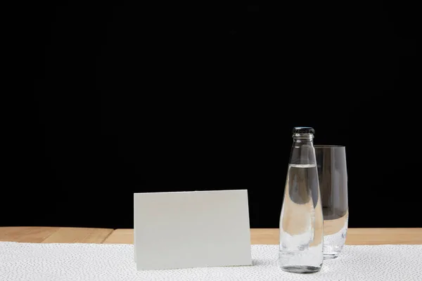 Botella con agua y vaso vacío en la mesa junto a la tarjeta en blanco sobre fondo negro - foto de stock