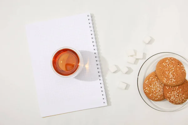 Top view of glass cup with fresh tea on blank notebook, sugar cubes and oatmeal cookies in plate on grey — Stock Photo