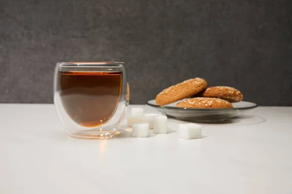 Vista de cerca del té caliente fresco en taza de vidrio, cubos de azúcar y galletas sabrosas en gris — Stock Photo