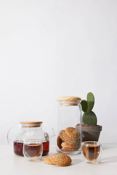 Té en tazas de vidrio, galletas sabrosas, tetera y cactus en olla sobre gris - foto de stock