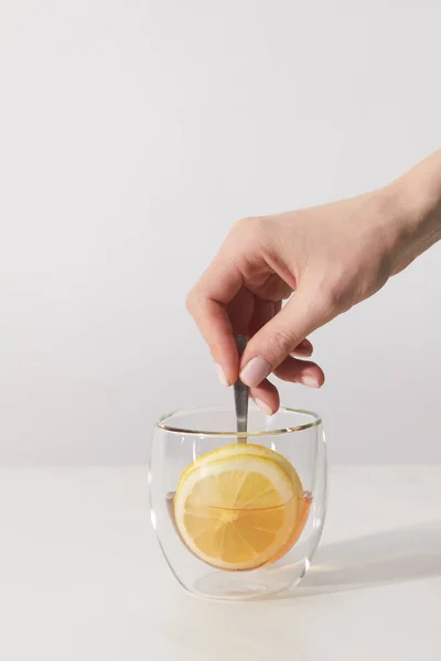 Plan recadré de main humaine et tasse en verre avec tisane fraîche, cuillère et tranche de citron sur gris — Photo de stock