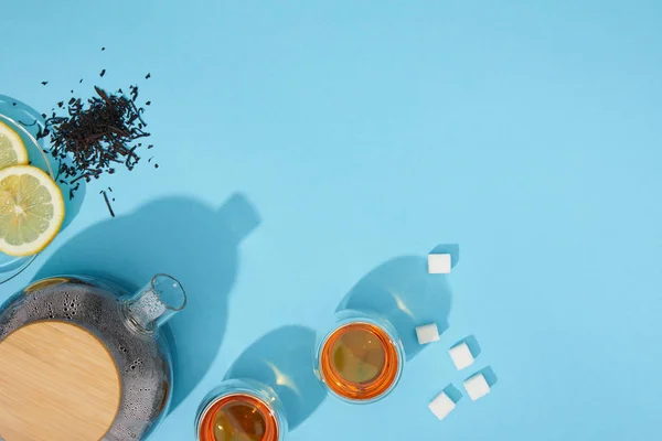 Top view of fresh tea in cups and teapot, sugar cubes, dry herbal tea and sliced lemon on blue — Stock Photo