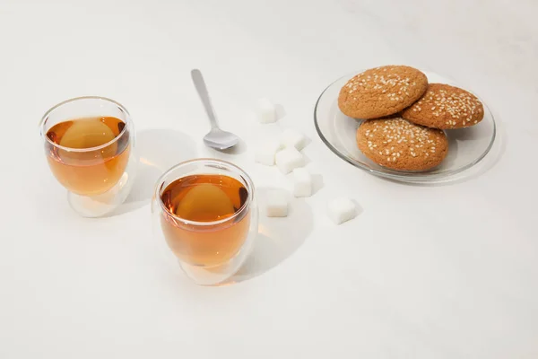 Vista de cerca del té en tazas de vidrio, cuchara, cubos de azúcar y galletas de avena en el plato en gris - foto de stock