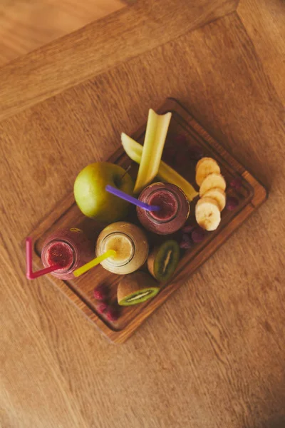 Raw fruits and bottles with smoothies on wooden board — Stock Photo