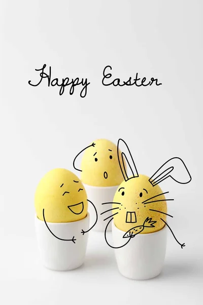 Три жовті розмальовані великодні яйця в яєчці стоїть з комічними намальованими обличчями на білому — стокове фото