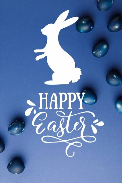 Vista superior de huevos de Pascua pintados de azul con letras de Pascua feliz y silueta de conejo en la superficie azul - foto de stock