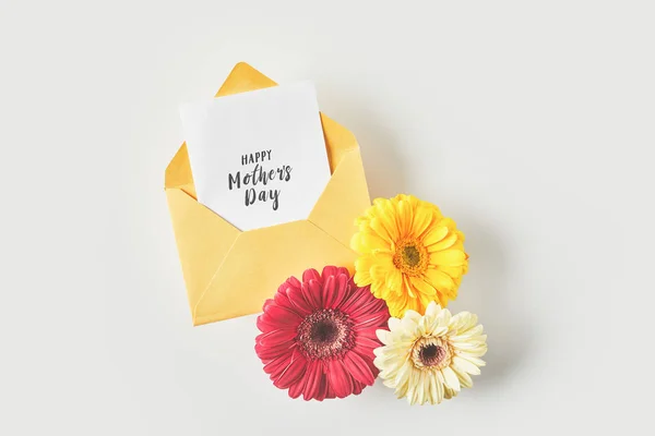 Top view of happy mothers day greeting card in envelope and beautiful gerbera flowers on grey — Stock Photo