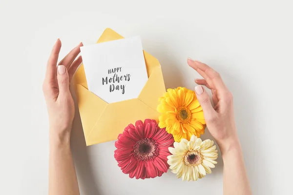Обрезанный снимок человеческих рук с конвертом, цветами герберы и поздравительной открыткой на день матери на сером — стоковое фото