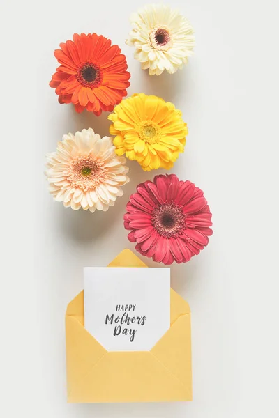 Vista superior de hermosas flores de gerberas tiernas y sobre con feliz tarjeta de felicitación del día de las madres en gris - foto de stock