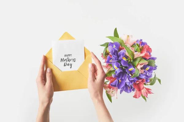 Colpo ritagliato di persona in possesso di busta con felice biglietto di auguri per la festa della mamma e bel mazzo di fiori su grigio — Foto stock