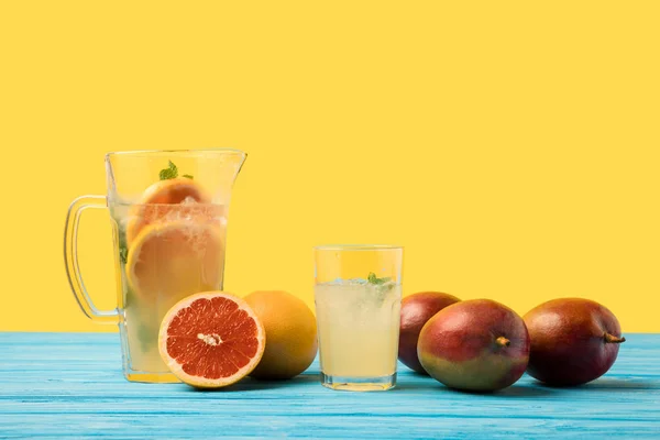 Свежие манго с грейпфрутами и холодным летним напитком в стакане и кувшине на бирюзовом деревянном столе — стоковое фото