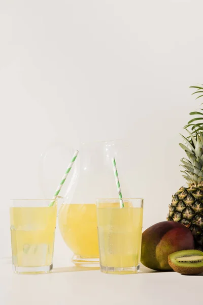 Glasses with straws and glass jug with juice and fresh tropical fruits on grey — Stock Photo