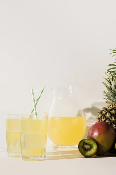 Vista de cerca de vasos y jarra de vidrio con jugo y frutas tropicales frescas en gris - foto de stock