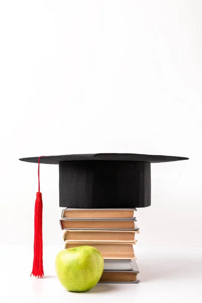 Apple near pile of books with academic cap on top isolated on white — Stock Photo
