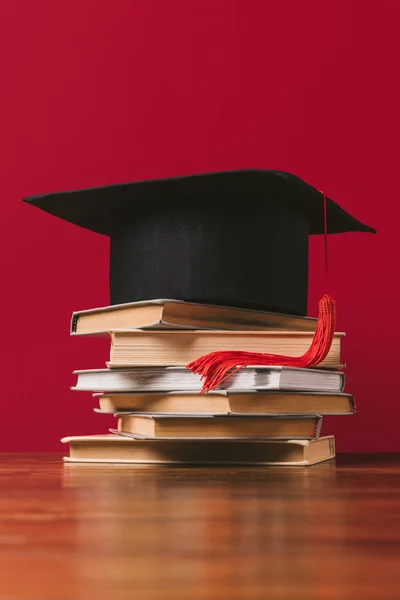 Academic cap on top of pile of books on red — Stock Photo