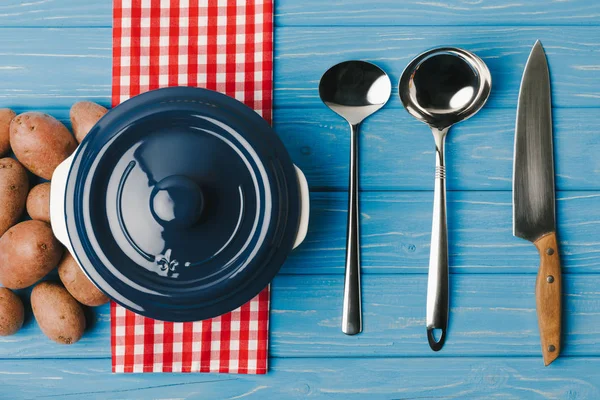 Top view of ladles, knife, pan and uncooked potatoes on blue table — Stock Photo