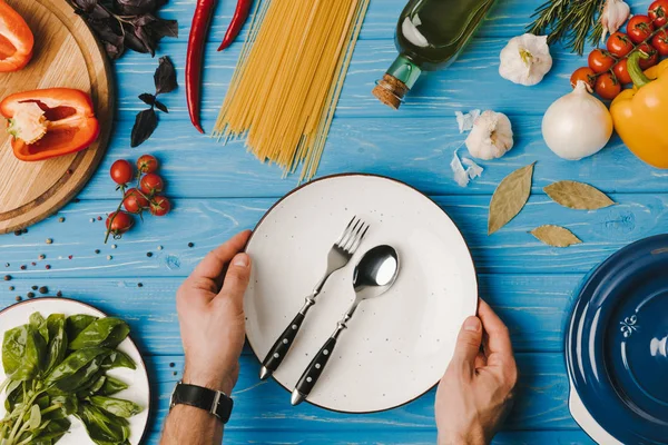 Imagen recortada del hombre poniendo placa con tenedor y cuchara en la mesa azul - foto de stock