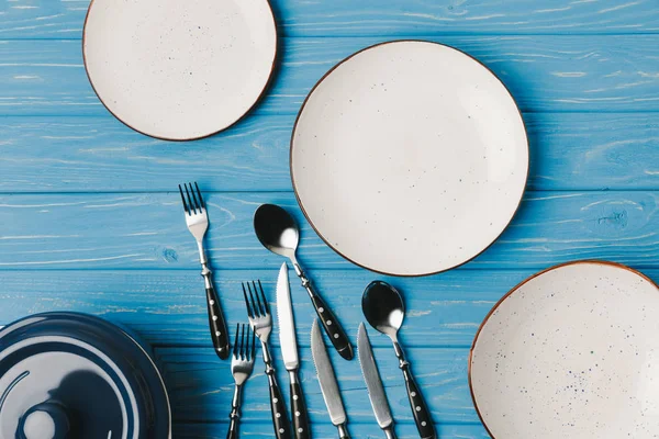 Vue de dessus des assiettes avec fourchettes, cuillères et couteaux éparpillés sur la table bleue — Photo de stock